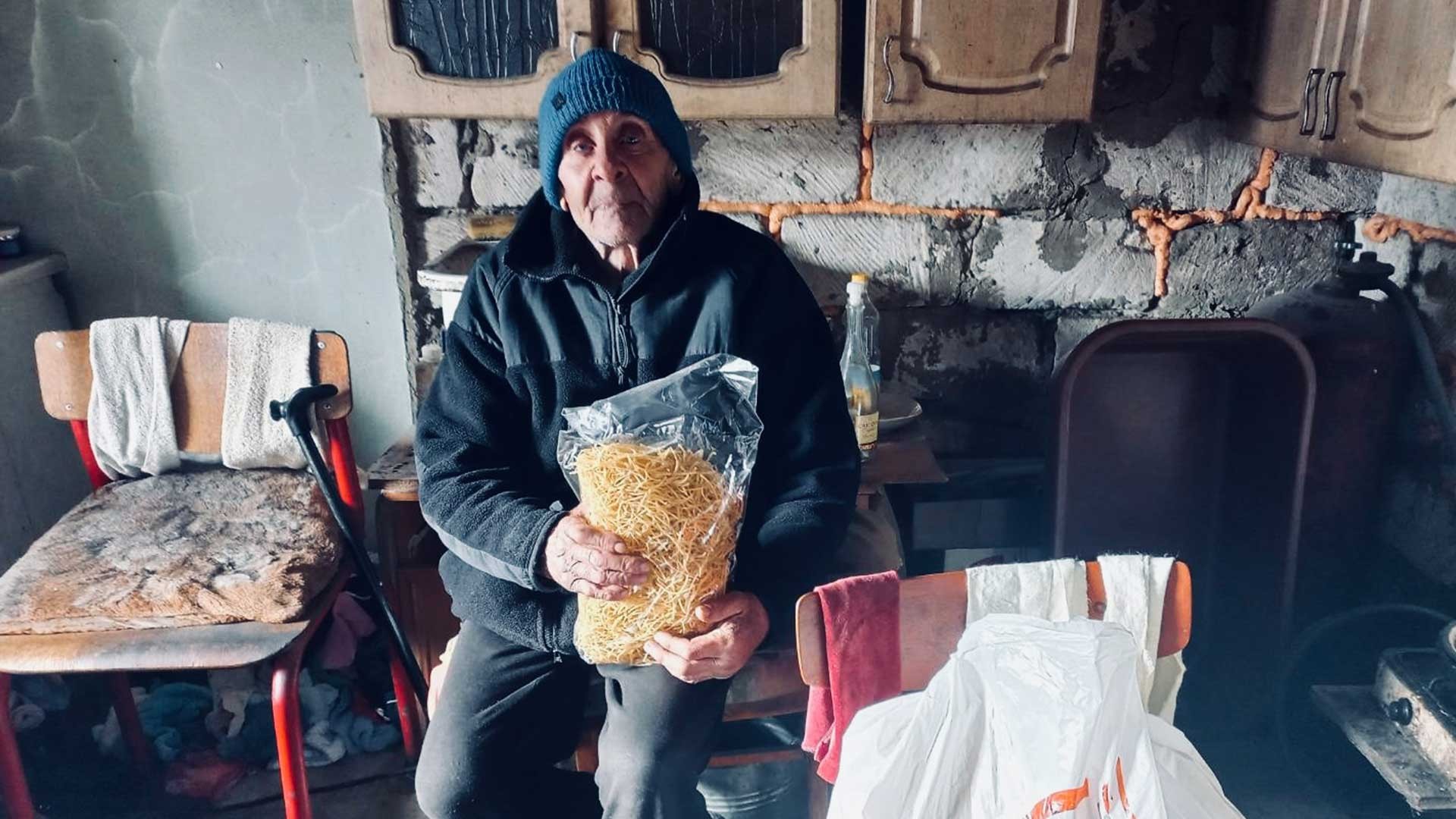 An elderly man in a draughty kitchen with a bag of supplies.