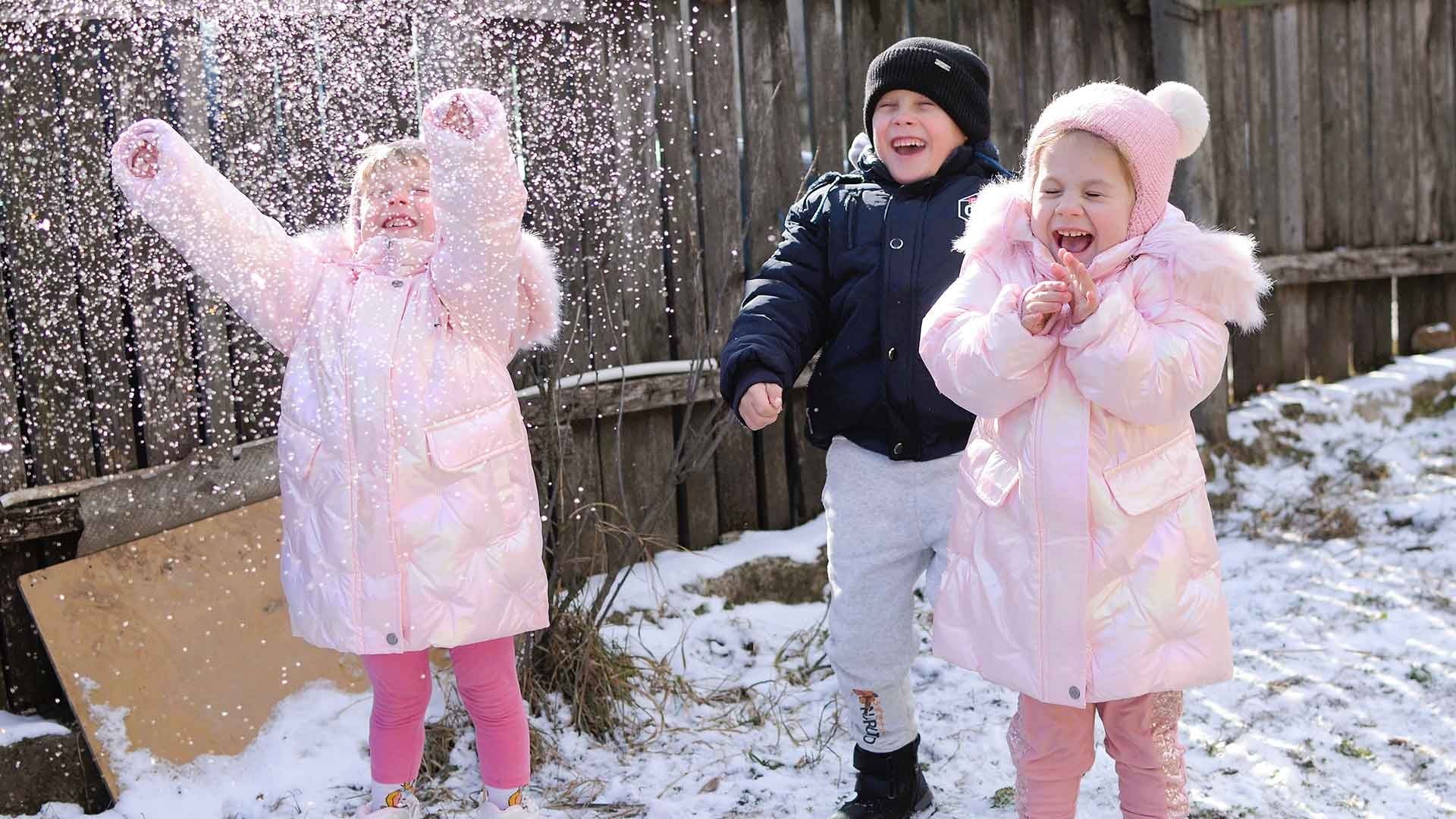 Three young children laugh and play together in the snow outdoors.